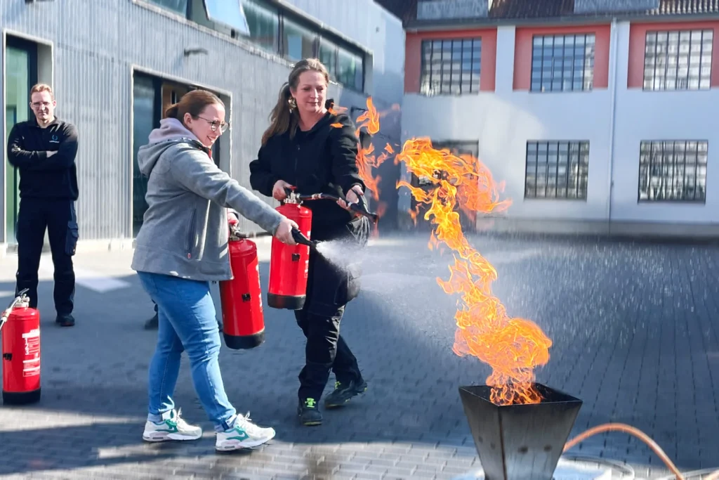 Zwei Frauen löschen mit Feuerlöschern eine brennende papiereimer-Attrappe auf dem Firmengelände